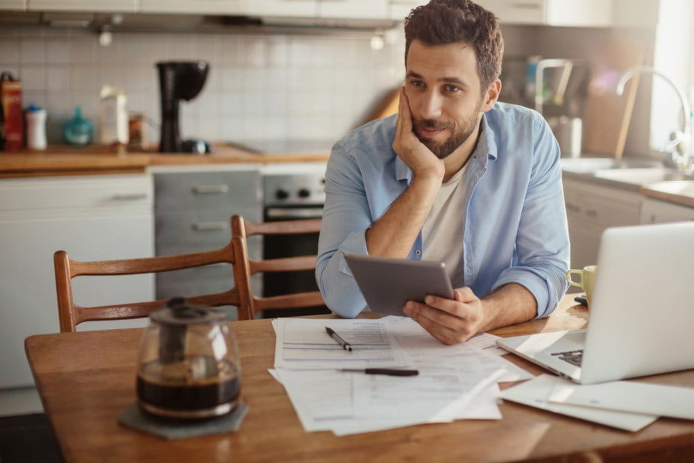 Smilende mann sitter og betaler regninger ved kjøkkenbordet